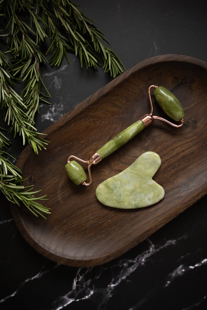 extra-programs-img From above of Jade roller and Gua Sha massage tool placed on wooden plate on black marble table with fresh twigs of rosemary